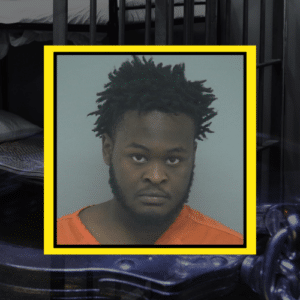 Mugshot of a black man with short dreadlocks, wearing an orange shirt.